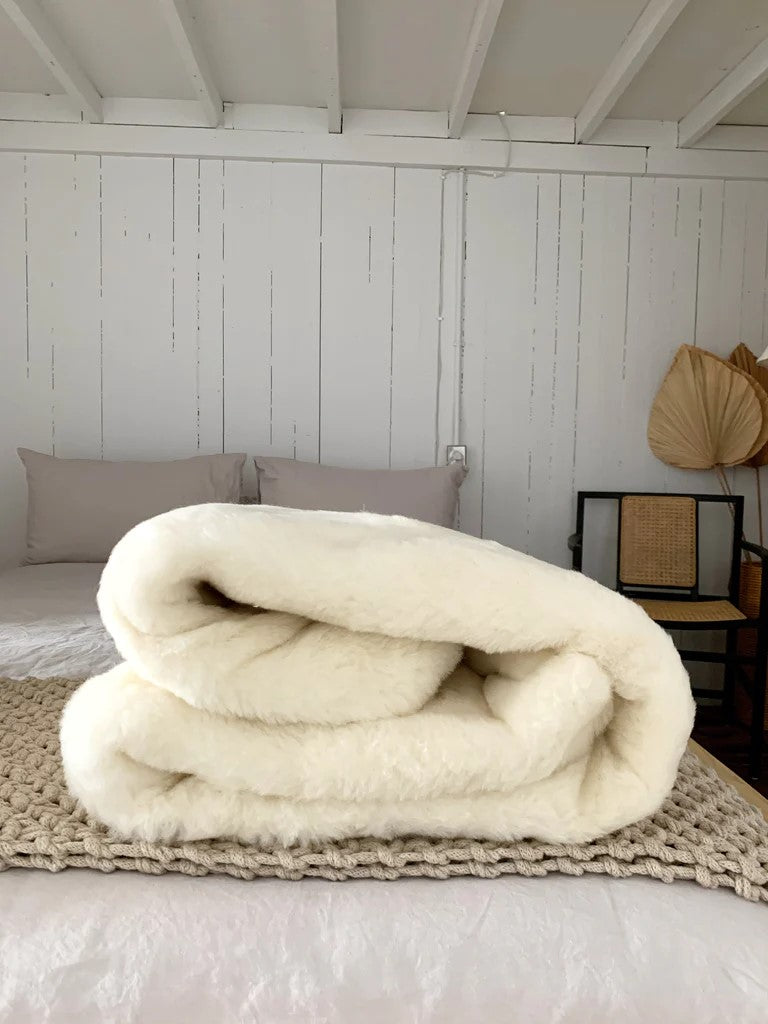 A folded white wool mattress overlay displayed on a bed with a wooden headboard in the background.